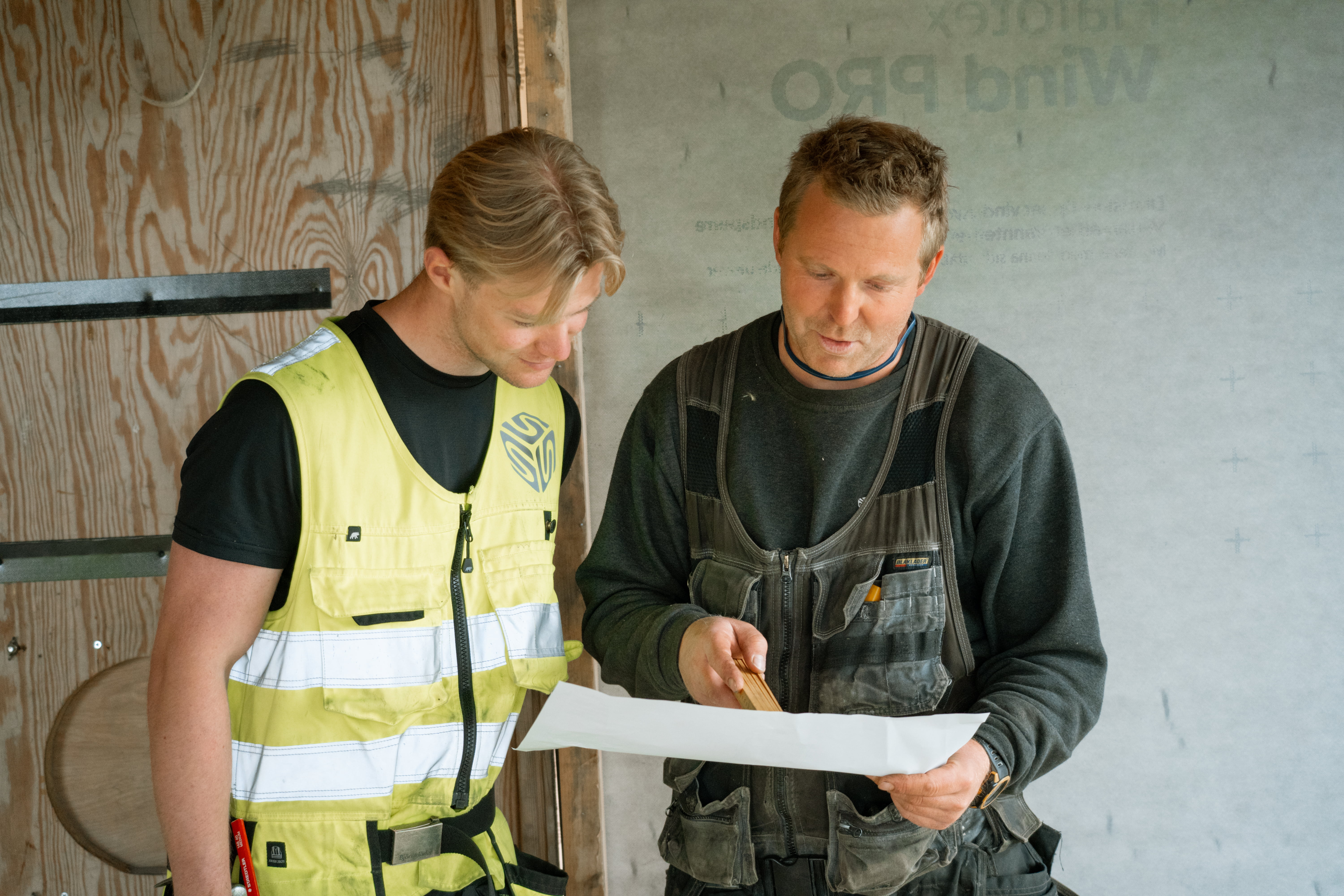 To personer i arbeidsklær ser på en byggetegning inne på en byggeplass med trevegger.