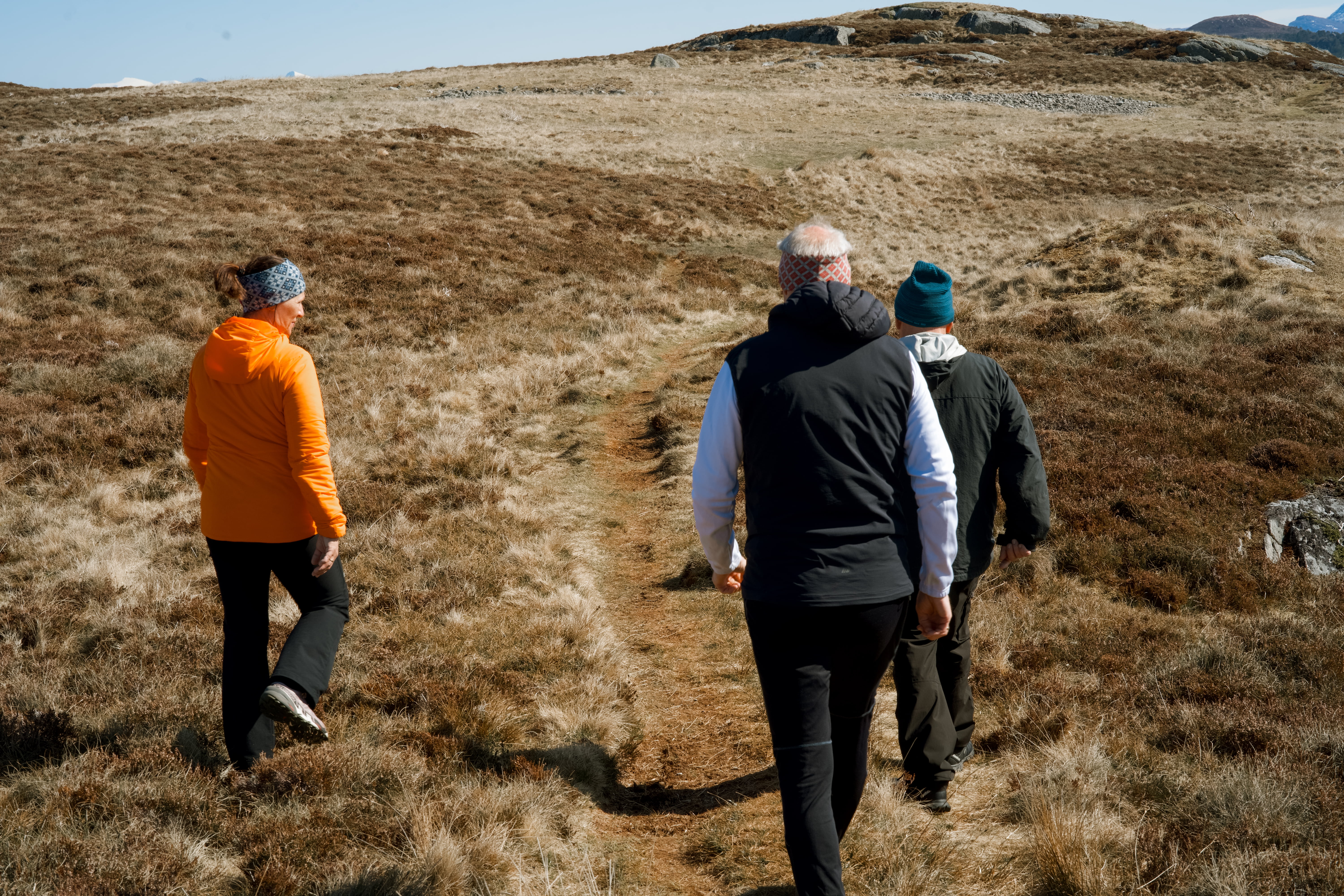 Tre personer går på en smal sti i et åpent landskap med gress og lyng.