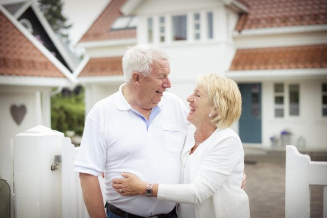 Eldre par holder rundt hverandre med hvit hus i bakgrunn. foto